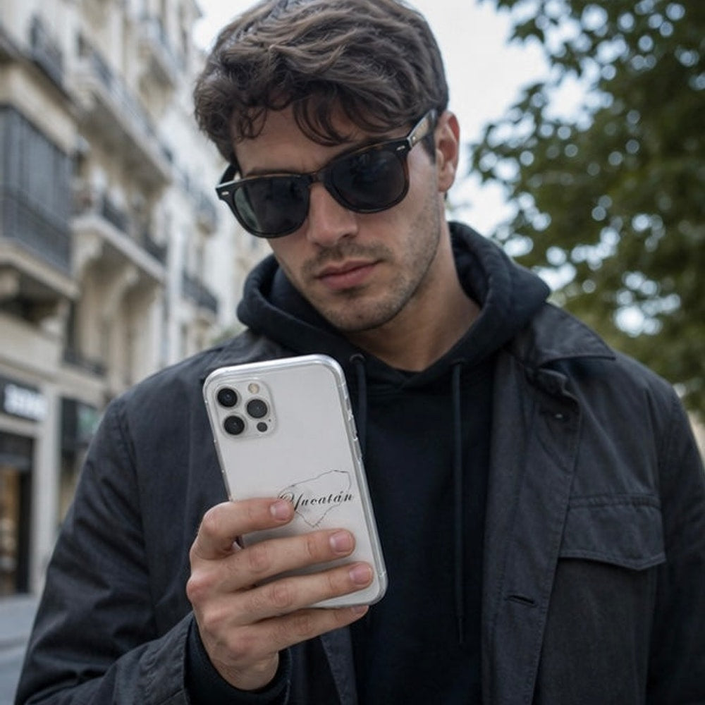 Man wearing sunglasses and a dark jacket holding a phone with a blurred urban background