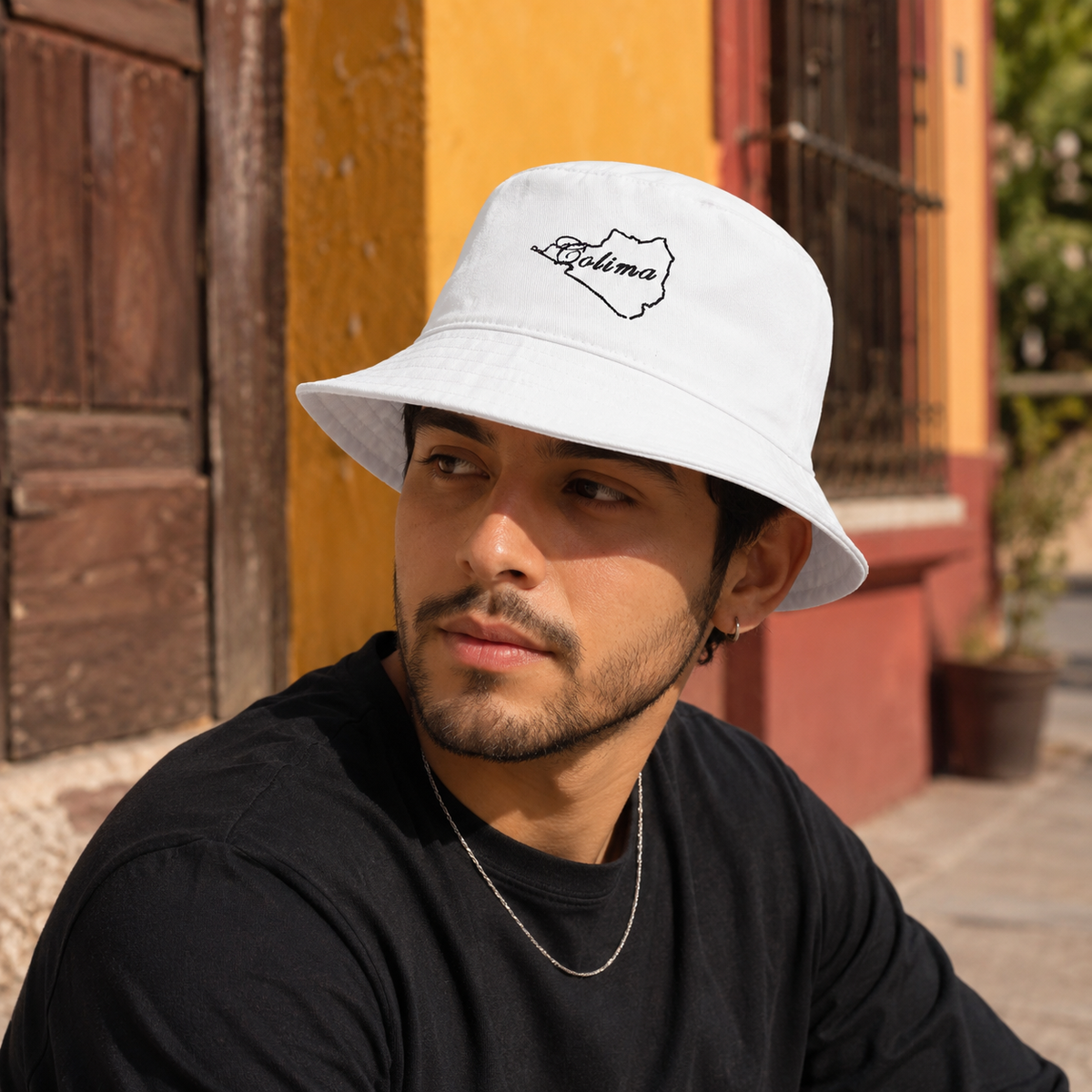 A man wearing a white bucket hat with a black script 'Colima' logo on the front, paired with a black t-shirt, photographed outdoors against a colorful yellow and red building background.