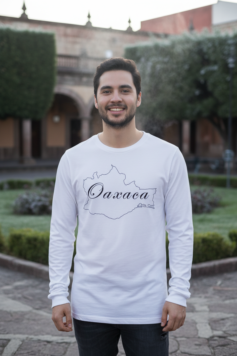 A smiling man wearing a white long-sleeve t-shirt with a black outline map of Oaxaca and the text 'Oaxaca' and 'Guatemala' printed on the front, standing outdoors in front of a historic building and green hedges.