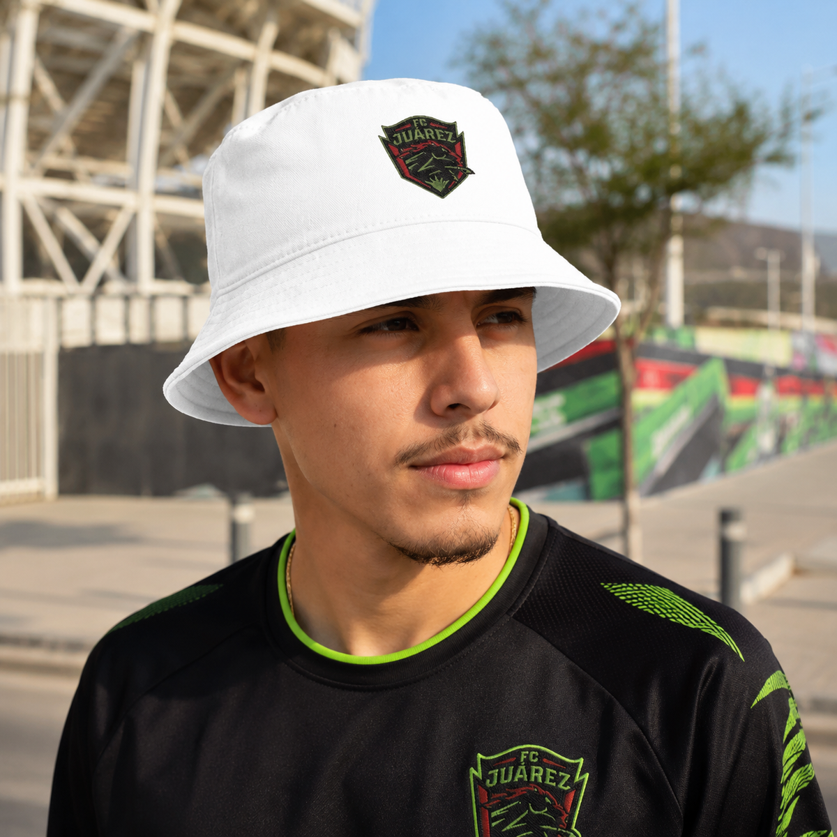 A man wearing a white bucket hat with a green and black FC Juárez crest, paired with a black athletic jersey featuring green accents and a matching crest, set against an outdoor stadium background.