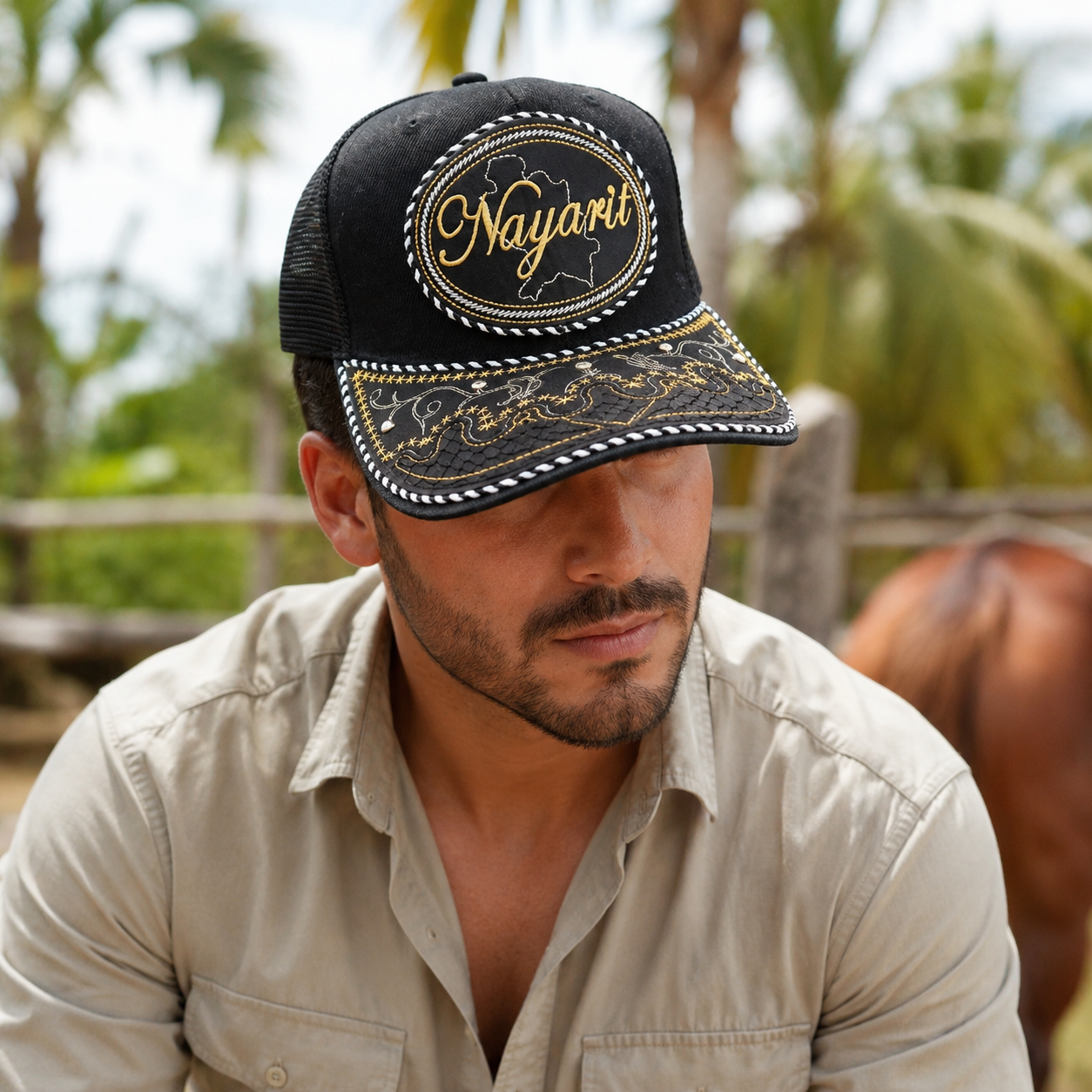 A Mexican model wearing a black Nayarit charro-style hat in an outdoor ranch setting with tropical vegetation. The embroidered Nayarit name and map design are clearly visible on the front.