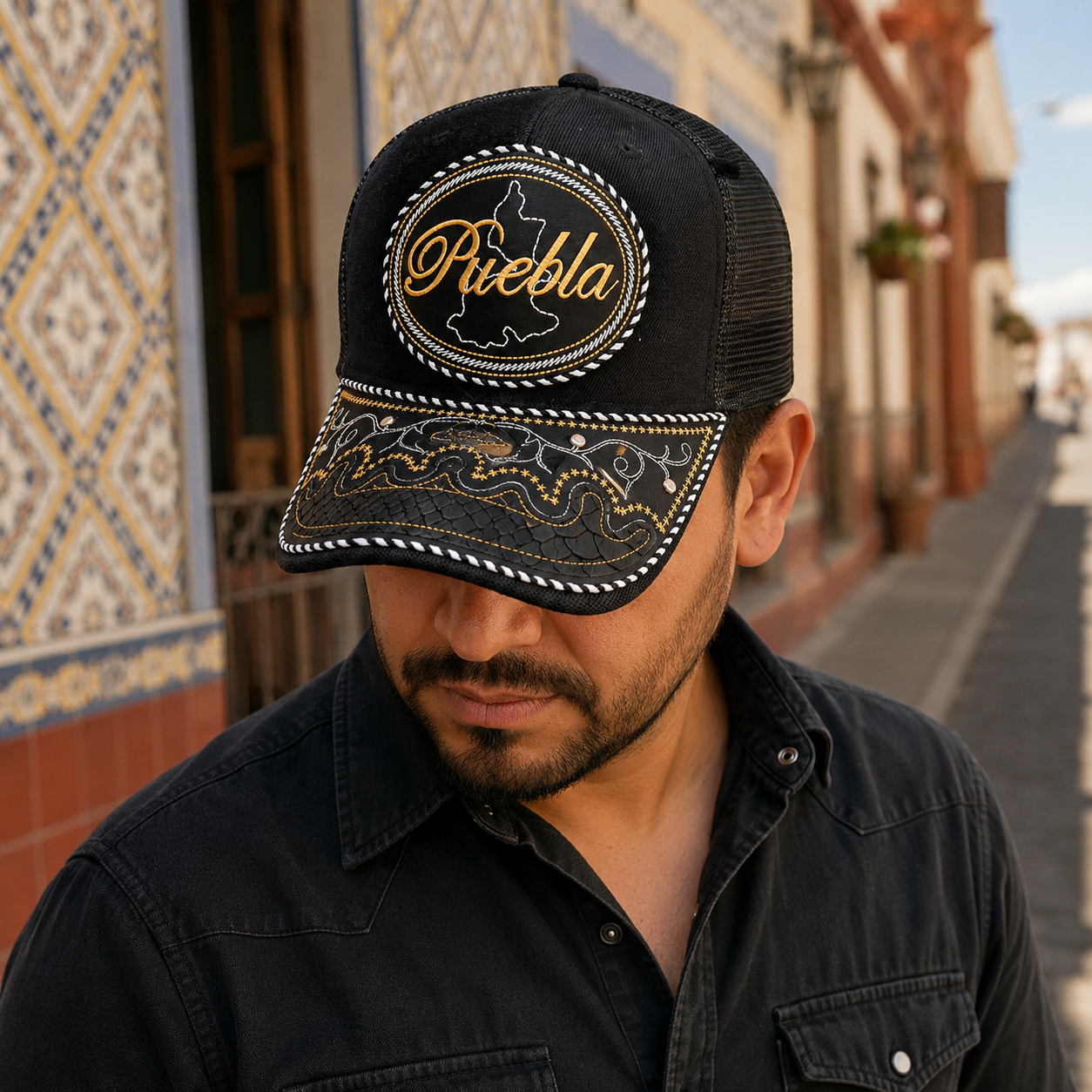A Mexican model wearing a black Puebla charro-style hat on a colonial street with tiled architecture. The embroidered Puebla name and map design are clearly visible on the front.