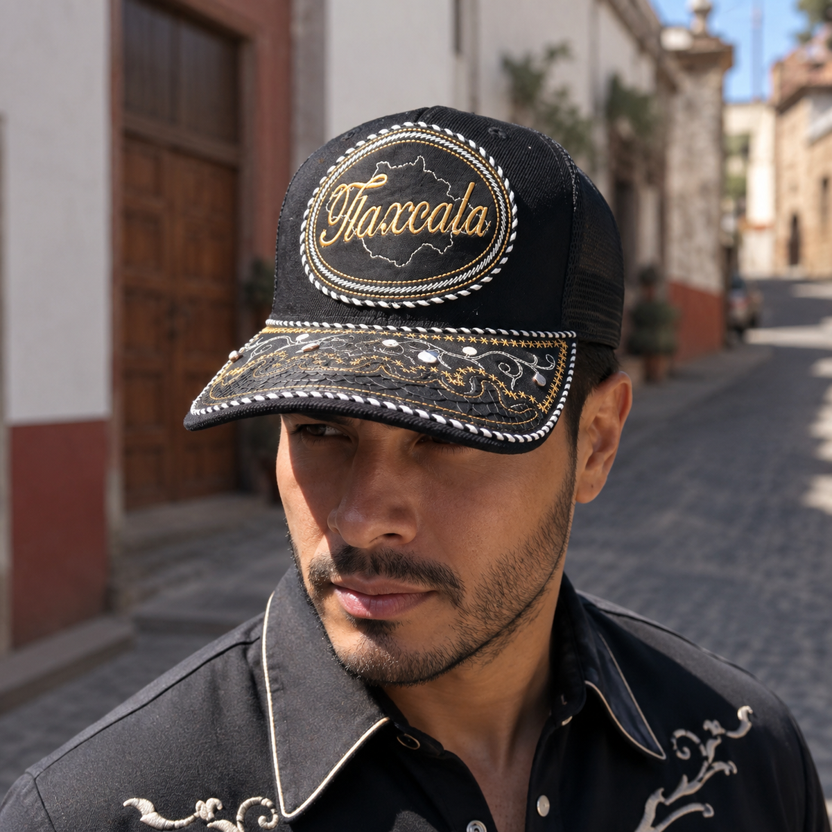 A Mexican model wearing a black Tlaxcala charro-style hat on a historic village street. The embroidered Tlaxcala name and map design are clearly visible on the front.