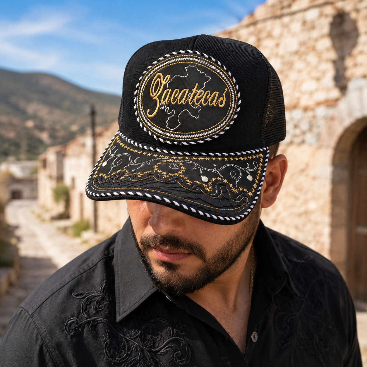 A Mexican model wearing a black Zacatecas charro-style hat in a rustic mountain town. The embroidered Zacatecas name and map design are clearly visible on the front. |