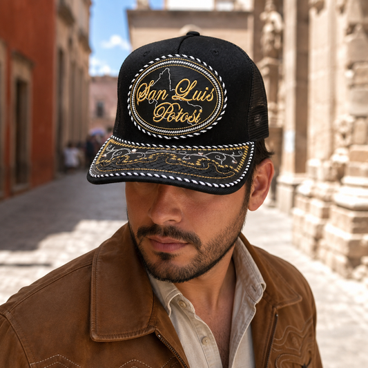 A Mexican model wearing a black San Luis Potosí charro-style hat on a colonial street with stone buildings. The embroidered San Luis Potosí name and map design are clearly visible on the front.
