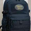 A black tactical backpack with a front zippered pocket and a circular embroidered patch reading 'Estado De Mexico' in gold, sitting on a wooden surface.