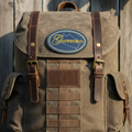 A vintage-style canvas backpack in brown and tan, featuring dark brown leather straps, brass hardware, and a blue oval 'Glencora' logo patch on the front, set against a rustic wooden background.