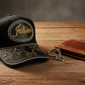 A black embroidered trucker hat with a curved brim and a brown leather wallet on a rustic wooden tabletop. The hat features a circular embroidered patch with the brand name 'Jalisco' in gold script, white stitched border details, and a black-and-gold patterned brim.