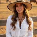 A woman wearing a wide-brimmed straw hat, white button-up shirt, and light blue jeans stands in front of a wooden fence in a sunlit field with horses in the background.