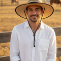 A smiling man wearing a wide-brim straw cowboy hat and a white button-up shirt stands in front of a wooden fence in a sunlit field, with horses visible in the background.