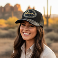 A smiling woman outdoors in a desert landscape at sunset wearing a black embroidered trucker hat with 'Sonora' in gold script and silver rhinestone trim, paired with a beige button-up shirt.