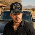 A man wearing a black trucker-style cap with gold embroidered details and the word 'Aguascalientes' on the front, photographed outdoors in front of a dark vehicle with a dry landscape background.