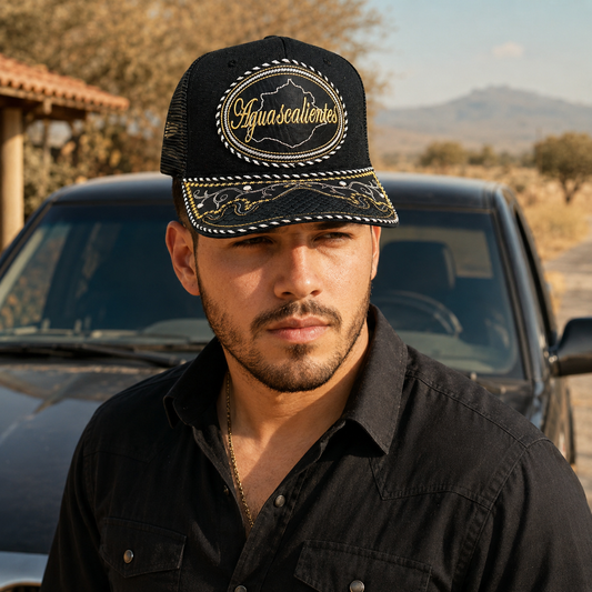 A man wearing a black trucker-style cap with gold embroidered details and the word 'Aguascalientes' on the front, photographed outdoors in front of a dark vehicle with a dry landscape background.