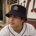 A man wearing a black bucket hat with a red and white embroidered 'Union Laguna' patch on the front, paired with a white baseball jersey with red pinstripes, set against a rustic outdoor background.