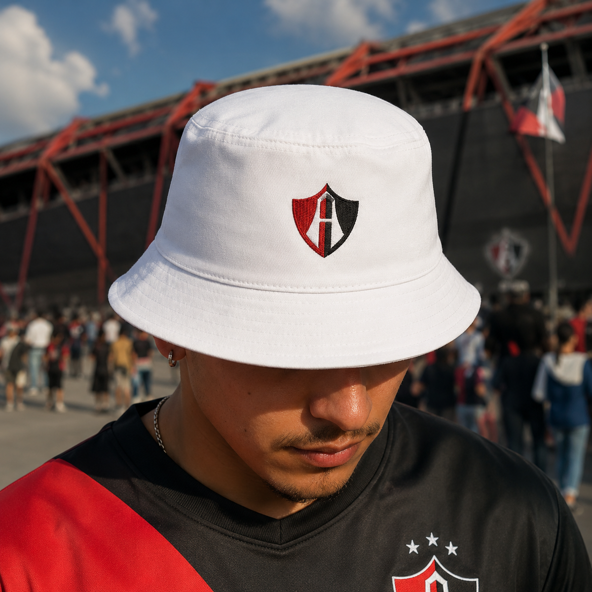 A man wearing a white bucket hat with a red and black Atlas shield logo, photographed outdoors in front of a stadium with people in the background.