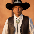 A man wearing a black cowboy hat with a silver decorative emblem, a white button-down shirt, a dark brown leather vest, and a silver bolo tie, standing against a warm, textured orange-brown wall.