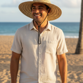 A man standing on a sandy beach wearing a wide-brimmed straw hat with a black chin cord, a light beige short-sleeve button-up shirt, and dark pants, with the ocean and palm trees in the background.