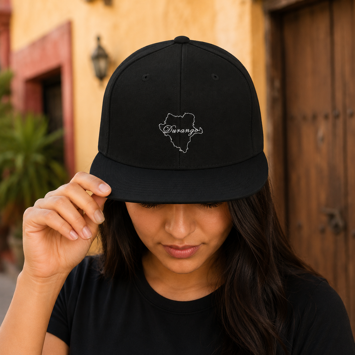 A woman wearing a black baseball cap with a white embroidered outline of the state of Durango and the word 'Durango' inside it, photographed outdoors against a warm-toned building background.