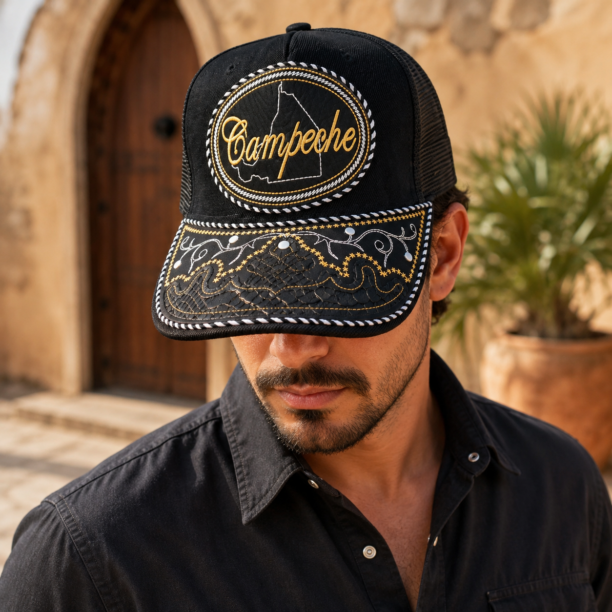 A Mexican model wearing a black Campeche charro-style hat outside a rustic hacienda. The embroidered Campeche name and map design are clearly visible on the front.