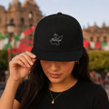 A woman wearing a black baseball cap with a white embroidered map of Jalisco and the word 'Jalisco' on the front, holding the brim with one hand in an outdoor setting with blurred buildings and flags in the background.