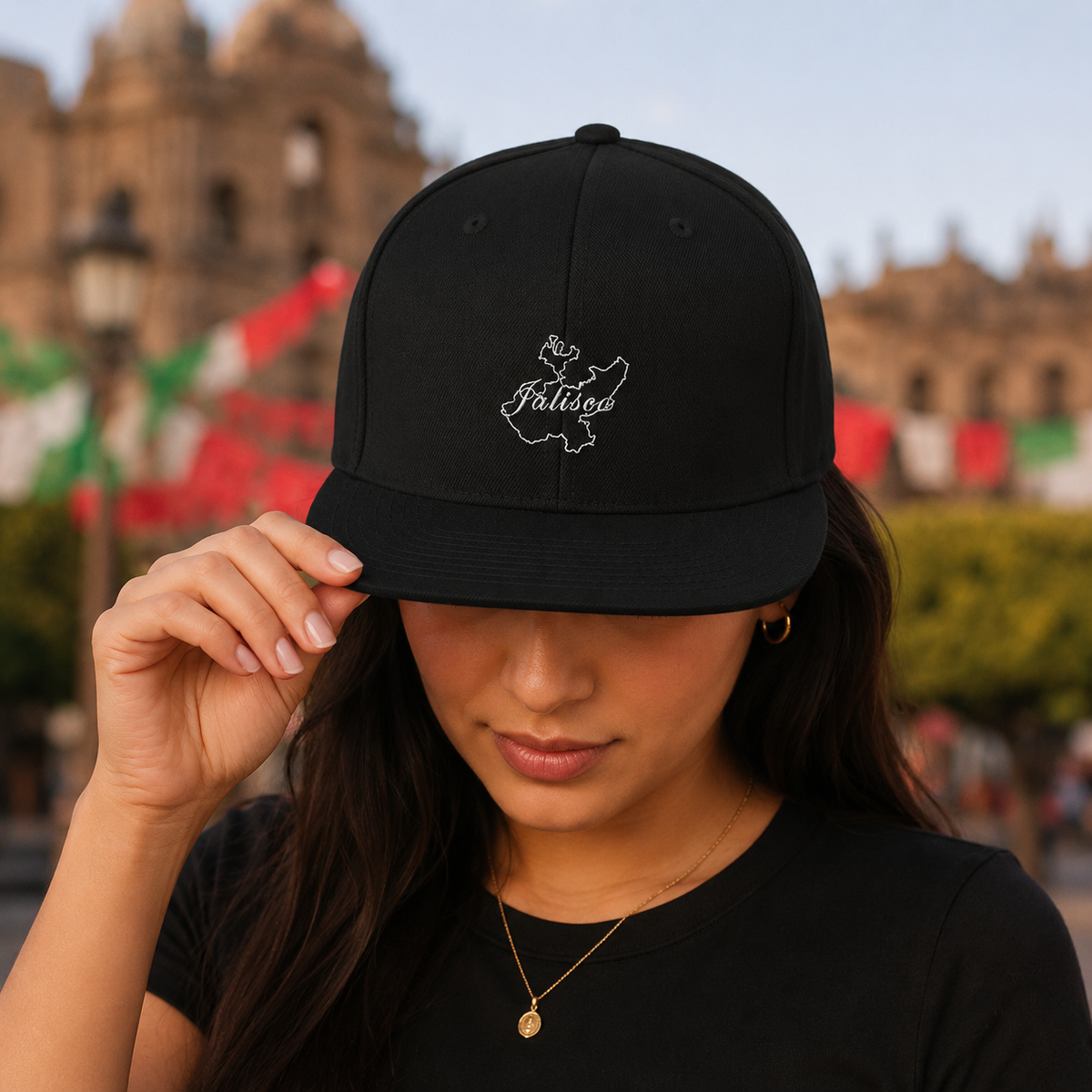 A woman wearing a black baseball cap with a white embroidered map of Jalisco and the word 'Jalisco' on the front, holding the brim with one hand in an outdoor setting with blurred buildings and flags in the background.