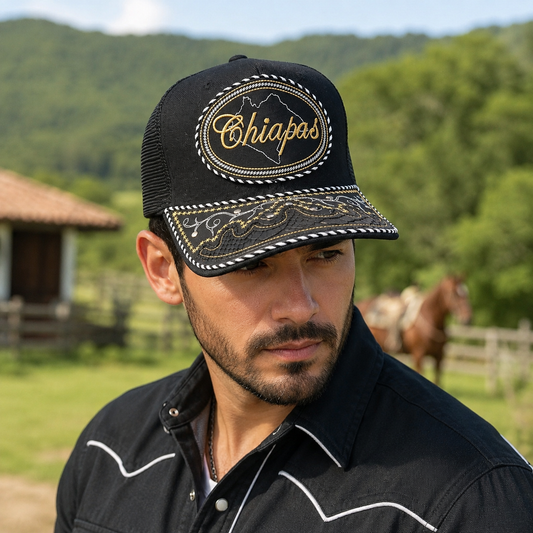 A Mexican model wearing a black Chiapas charro-style hat in green countryside near a traditional ranch. The embroidered Chiapas name and map design are clearly visible on the front.