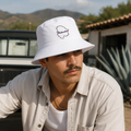 A Mexican model wearing the Chihuahua bucket hat in a realistic outdoor lifestyle setting, with the front design clearly visible. The Chihuahua bucket hat design, shape, and colors are shown clearly in a natural casual scene.