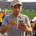 A man in a stadium wearing a gray t-shirt and beige cap takes a photo of a white smartphone with a blue and yellow Tigres jersey design on the back.