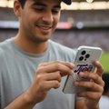 A man in a gray t-shirt and navy baseball cap holds a white smartphone with a 'Tigers' baseball case in a stadium setting.