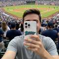 A man taking a selfie in a crowded baseball stadium, holding a white phone on a  'Bravos' case. The field is visible in the background with fans seated in the stands.