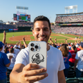 A man taking a selfie at a baseball stadium, holding a white phone with a black 'Guerreros' logo. He is wearing a white t-shirt, with the green field, crowd, and blue sky in the background.