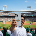 A man in a white t-shirt at a baseball stadium takes a selfie with a silver smartphone featuring a green and gold Leones de Yucatan logo on the back. The stadium is filled with fans, and players are visible on the field under a clear blue sky.