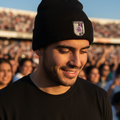 A man wearing a black knit beanie with a Queretaro crest patch on the front, photographed in a stadium crowd at sunset.