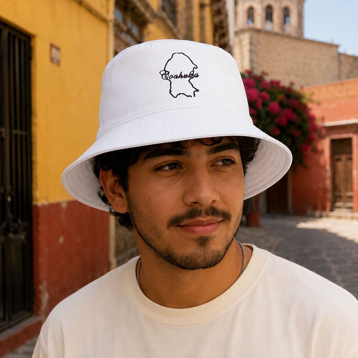 A man wearing a white bucket hat with a black outline graphic of the state of Coahuila and the word 'Coahuila' written inside, set against a warm, sunlit outdoor street background.
