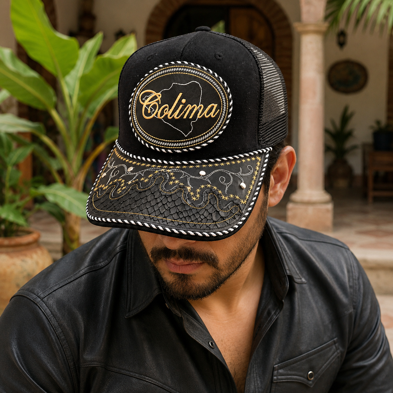A Mexican model wearing a black Colima charro-style hat in an outdoor hacienda patio with tropical greenery. The embroidered Colima name and map design are clearly visible on the front.