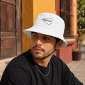 A man wearing a white bucket hat with a black script 'Colima' logo on the front, paired with a black t-shirt, photographed outdoors against a colorful yellow and red building background.