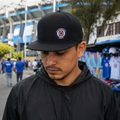 A Mexican model wearing the Cruz Azul snapback hat in a realistic outdoor lifestyle setting, with the front design clearly visible. The Cruz Azul snapback hat design, shape, and colors are shown clearly in a natural casual scene.