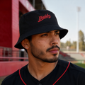 A Mexican model wearing the Diablos Rojos del Mexico bucket hat in a realistic outdoor lifestyle setting, with the front design clearly visible. The Diablos Rojos del Mexico bucket hat is shown naturally.