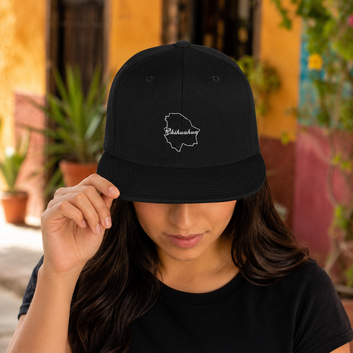 A woman wearing a black baseball cap with a white embroidered outline of Chihuahua and the word 'Chihuahua' on the front, holding the brim with one hand in an outdoor setting.