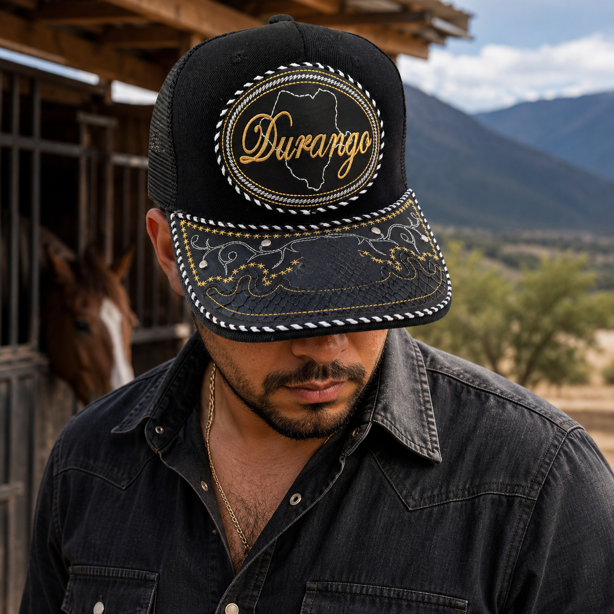 A Mexican model wearing a black Durango charro-style hat near a rustic stable and mountain landscape. The embroidered Durango name and map design are clearly visible on the front.