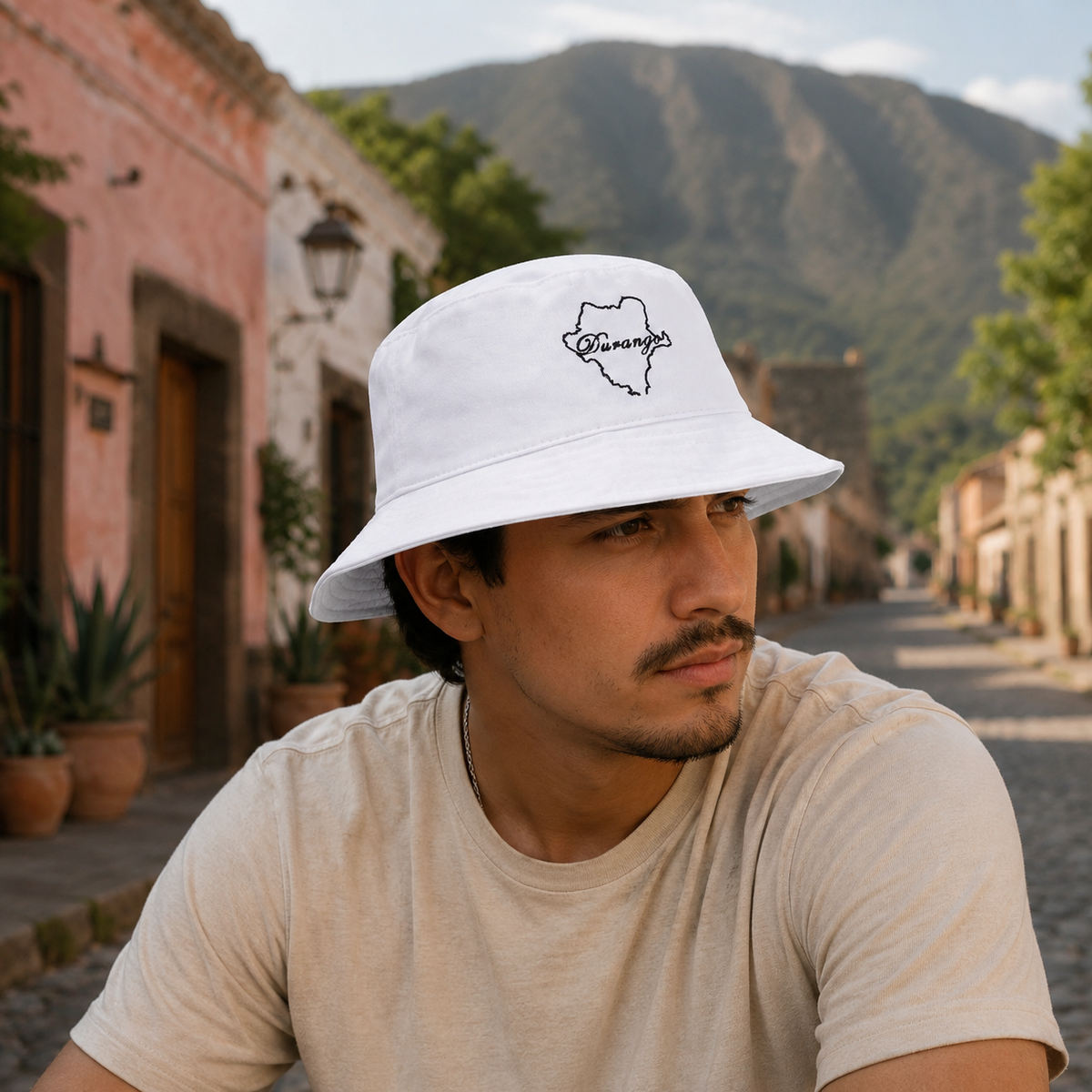A man wearing a white bucket hat with a black outline map graphic and the word 'Durango' on the front, paired with a beige t-shirt, sitting on a cobblestone street with a mountain and buildings in the background.