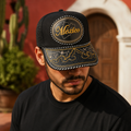 A man wearing a black trucker-style cap with gold embroidered details, including a map outline and the word 'México' on the front panel, photographed outdoors in front of a warm-toned building.