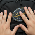 A close-up of a black backpack with an oval embroidered patch reading 'Ayuascalientes' in yellow script. Two hands are visible: one holding the patch in place,.