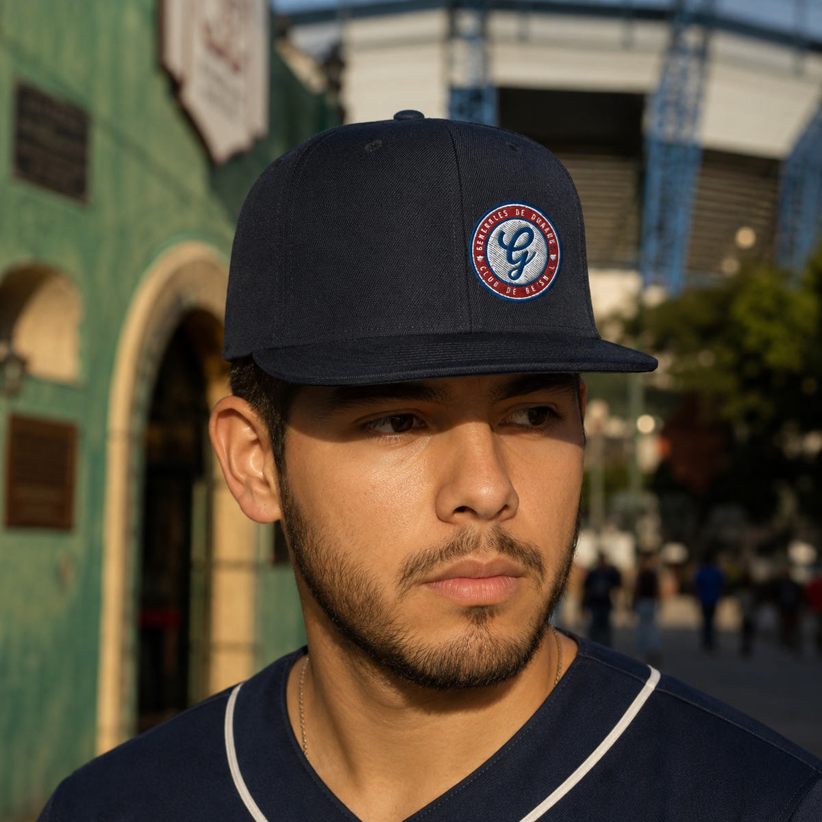A man wearing a navy blue baseball cap with a circular 'Generales de Durango' logo patch on the front, photographed outdoors in warm sunlight with a blurred stadium and building background.