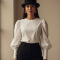 A woman wearing a white long-sleeve blouse with puffed balloon sleeves, black trousers, a black bowler hat, and gold hoop earrings, standing against a warm beige wall.