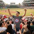 A man in a crowded baseball stadium stands with his arms raised, wearing a gray t-shirt with a red, white, and blue Generales de Durango logo. Fans and popcorn buckets are visible in the foreground, with the baseball field and stadium stands in the background.