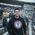 A man smiling in a crowded stadium wearing a black hoodie with a circular red, white, and blue Generales de Durango  logo on the chest, paired with blue jeans.