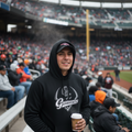 A man wearing a black hoodie with a white 'Guerreros' graphic stands in a crowded baseball stadium, holding a takeaway coffee cup.