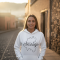 A woman stands on a cobblestone street wearing a white hoodie with a black outline map graphic and the word 'Hidalgo' printed across the chest. The background shows warm, sunlit buildings and a softly blurred outdoor setting.