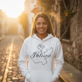 A smiling woman stands on a sunlit cobblestone street wearing a white pullover hoodie with a black outline map graphic and the word 'Jalisco' across the chest, paired with black pants.